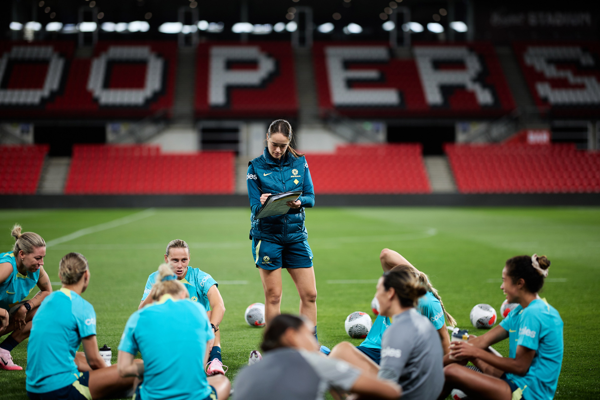 A woman with a clipboard is surrounded by a circle of seated soccer players on the field