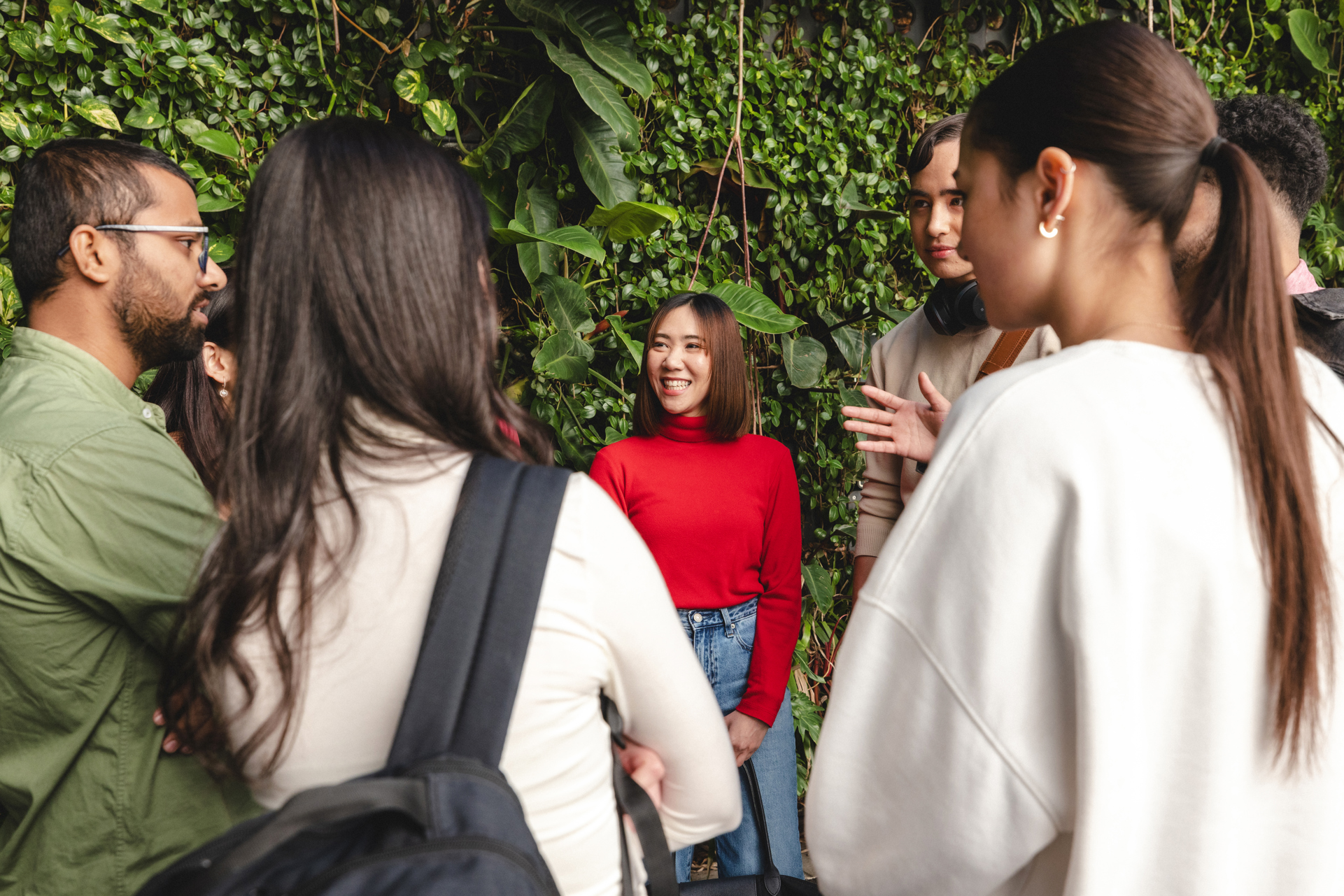 Male and female students standing chatting in a circle with green plant wall behind them.