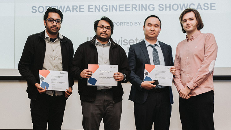 Four men proudly display certificates in front of a large screen, smiling and celebrating their achievements.